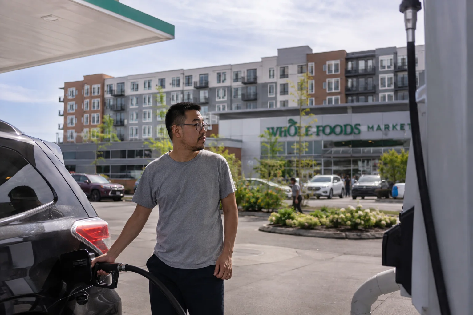 Man pumping gas into a car at a gas station, with a supermarket and residential buildings in the background in the United States.