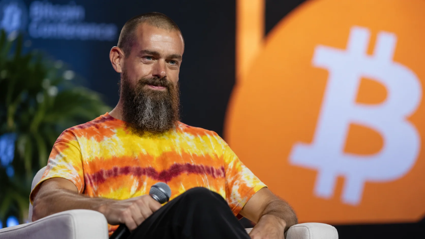 Jack Dorsey seated on a panel with a microphone, in front of a large Bitcoin symbol on an orange background.