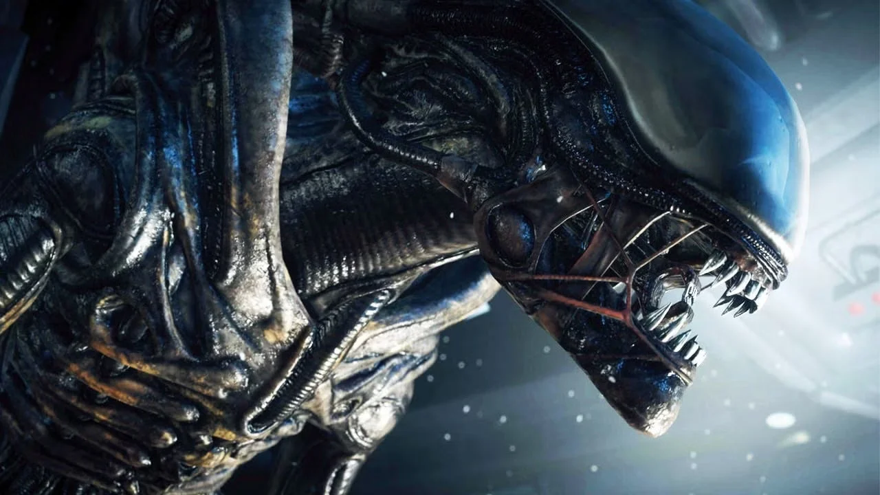 Close-up of a biomechanical-looking xenomorph, with an elongated head, open jaw, and exposed teeth, lit by cold light in a science fiction environment.