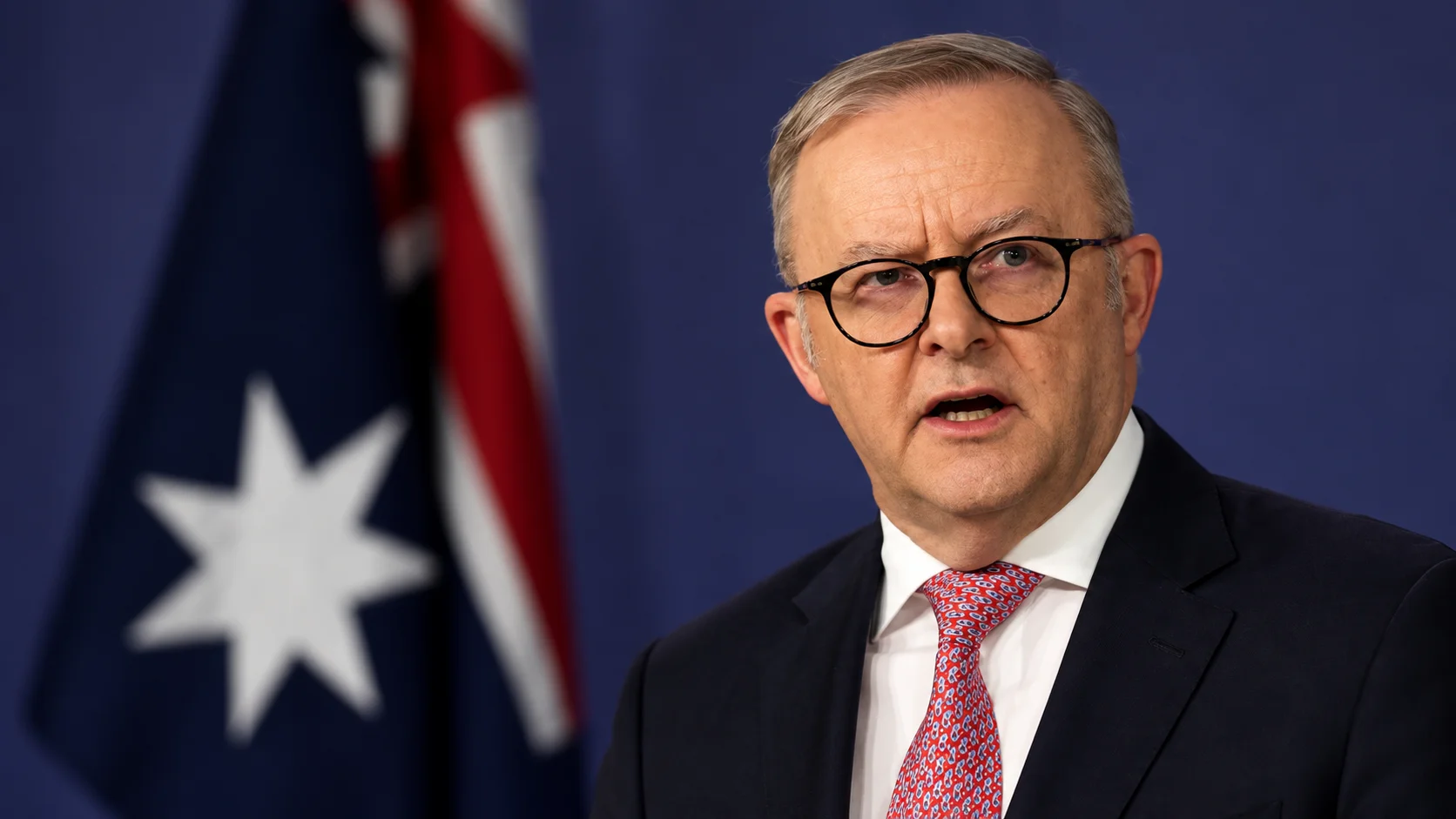 Primer plano de un funcionario australiano hablando, con una bandera australiana desenfocada de fondo.