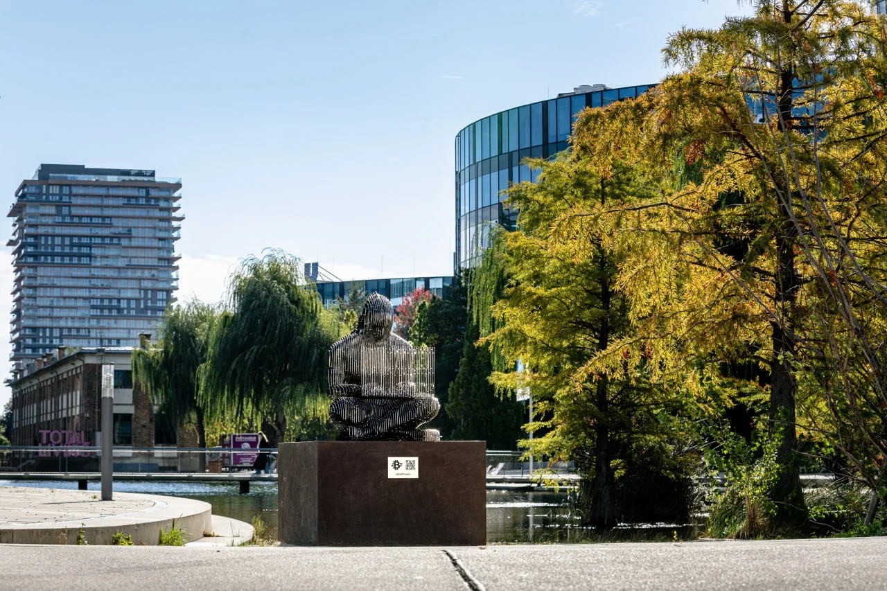 Escultura de Satoshi Nakamoto sentado com um laptop sobre um pedestal à beira de um lago em Viena, cercada por árvores e edifícios modernos. A figura é formada por finas camadas de aço inoxidável.