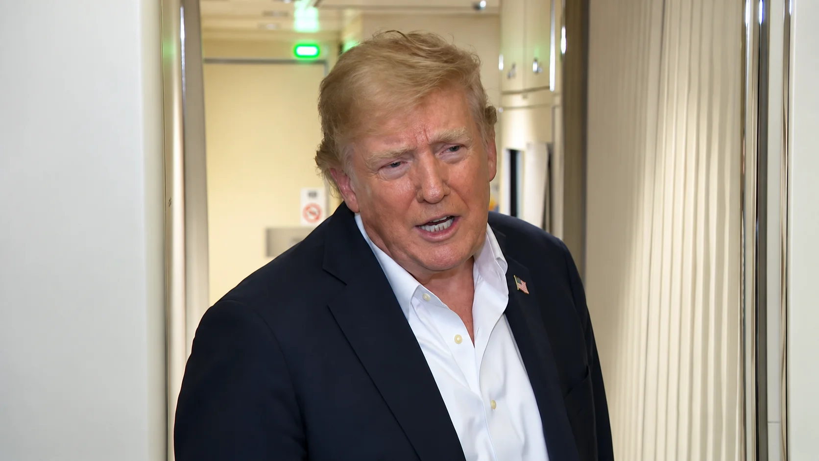 Donald Trump speaks with journalists in an aircraft aisle, wearing a dark suit and white shirt.