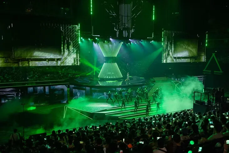 Arena de esports com palco iluminado em verde, efeitos de luz, fumaça e público acompanhando o evento.